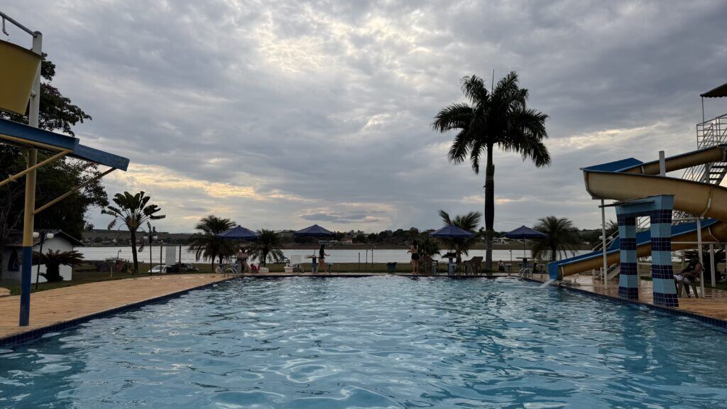 Piscina grande do clube ao entardecer, com palmeiras e guarda-sóis