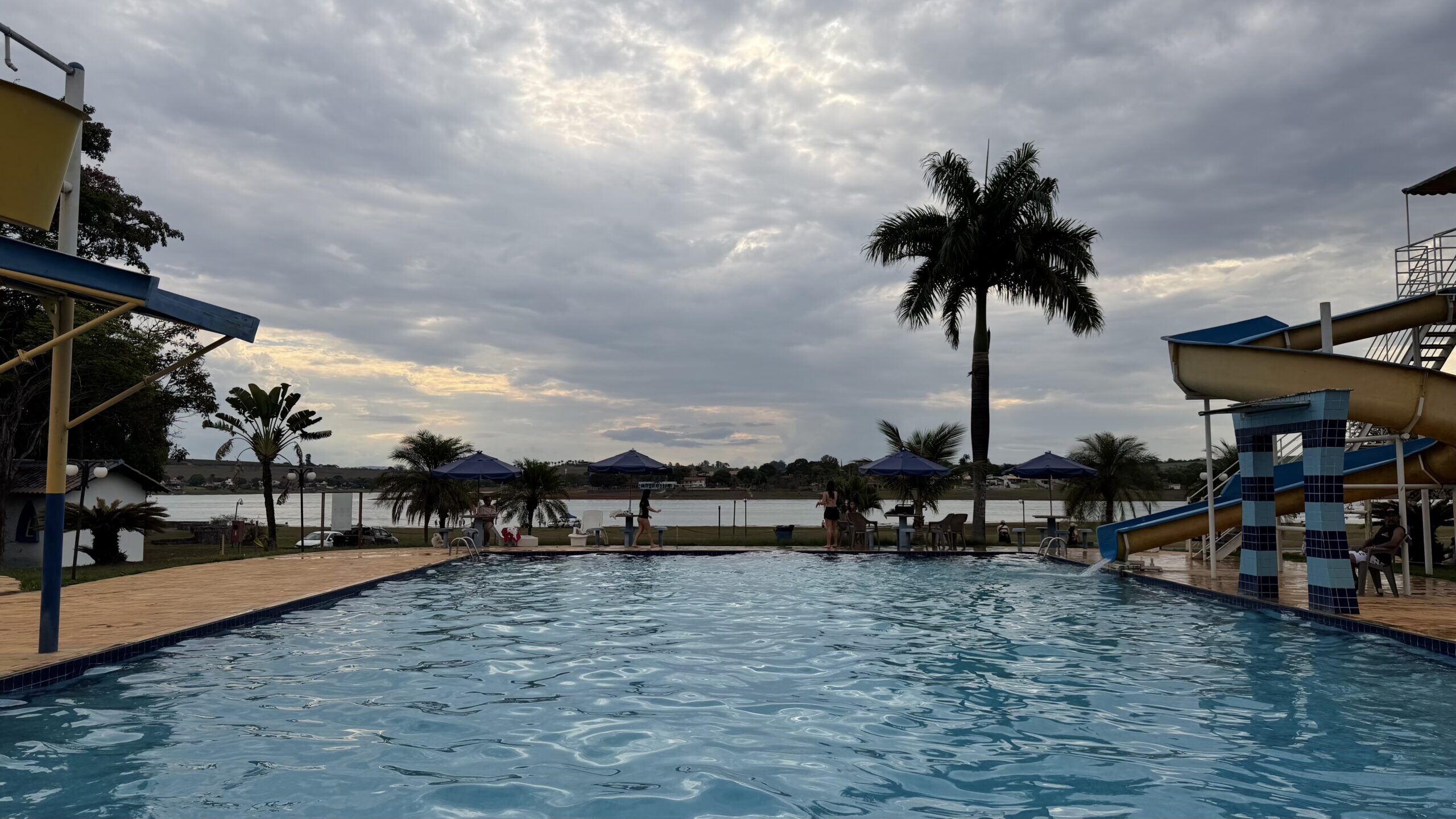 Piscina grande do clube ao entardecer, com palmeiras e guarda-sóis